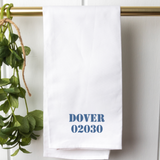 A white cotton tea towel with the text 'DOVER 02030' printed in blue ink, displayed on a banister alongside a green plant. Available at Be Charmed