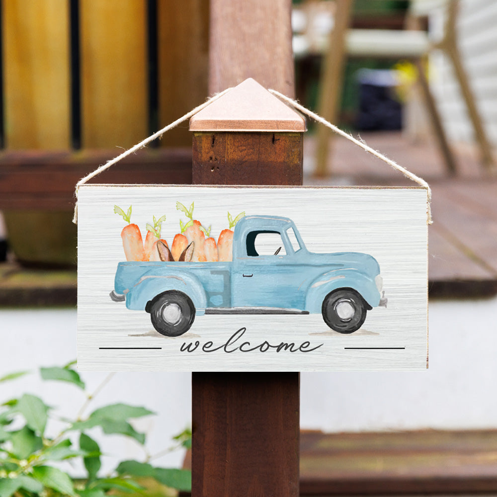 A rustic wooden twine sign featuring an illustration of a blue truck with carrots in the back, and the text 'Welcome' on a wooden post. Available at Be Charmed