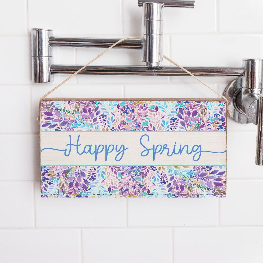 A rustic wooden sign with a floral pattern and the phrase 'HAPPY SPRING' written in bold blue letters, attached to a bathroom wall. Available at Be Charmed