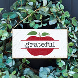 A rustic wooden sign with a split apple design and the word 'grateful' written on it, hung by twine with green leaves in the background. Available at Be Charmed
