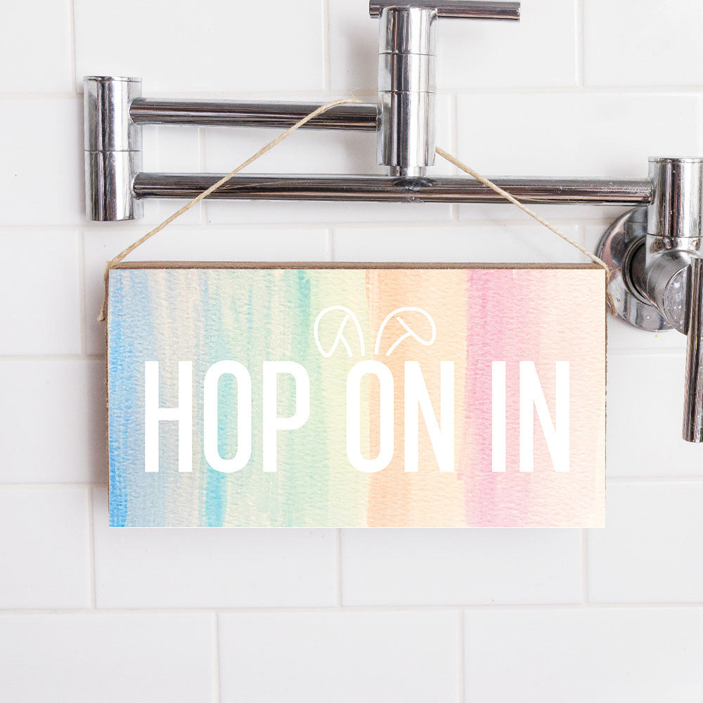 A wooden twine sign with a multicolored background and the text 'HOP ON IN' displayed, hanging on a wall with a shower in the background. Available at Be Charmed