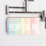 A wooden twine sign with a multicolored background and the text 'HOP ON IN' displayed, hanging on a wall with a shower in the background. Available at Be Charmed