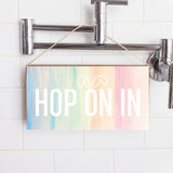 A wooden twine sign with a multicolored background and the text 'HOP ON IN' displayed, hanging on a wall with a shower in the background. Available at Be Charmed