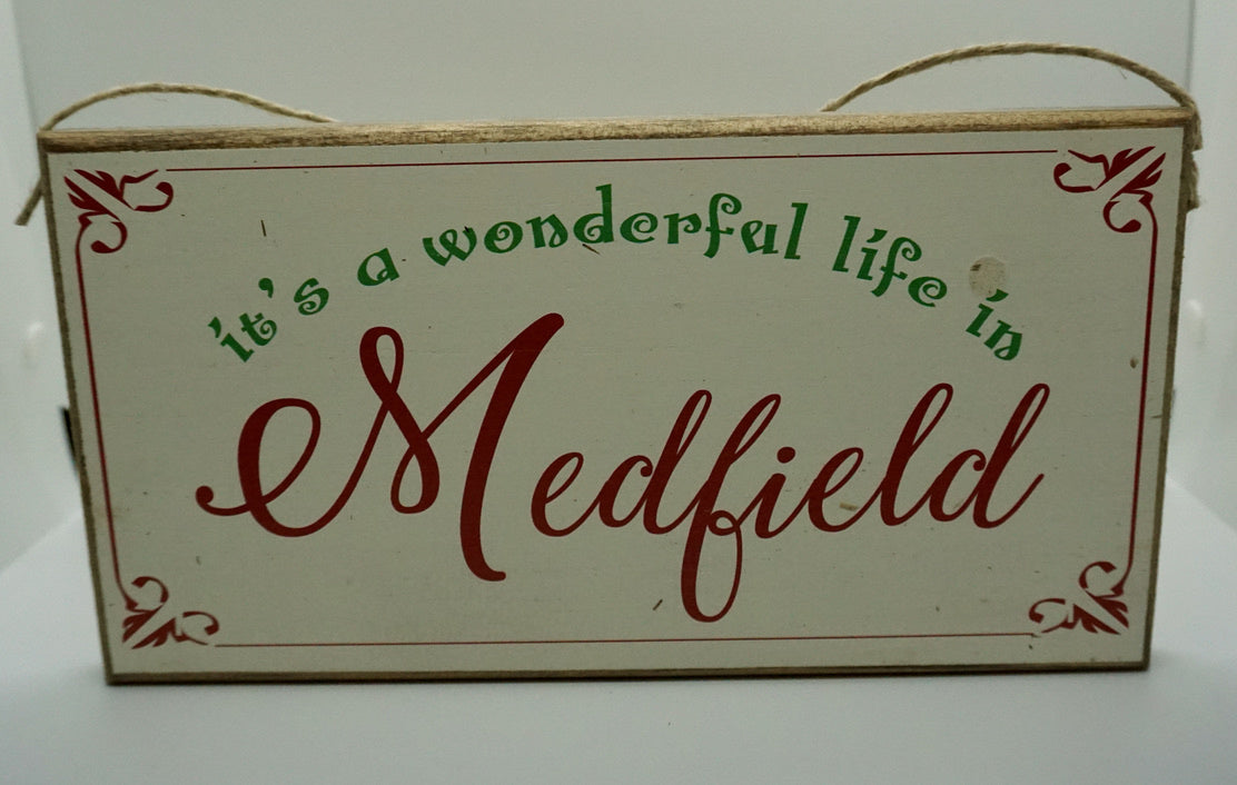 A rustic wooden twine sign with the text 'It's A Wonderful Life in Medfield' written in a decorative font, surrounded by a floral border and tied with twine at the top. Available at Be Charmed