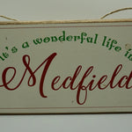 A rustic wooden twine sign with the text 'It's A Wonderful Life in Medfield' written in a decorative font, surrounded by a floral border and tied with twine at the top. Available at Be Charmed