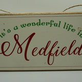 A rustic wooden twine sign with the text 'It's A Wonderful Life in Medfield' written in a decorative font, surrounded by a floral border and tied with twine at the top. Available at Be Charmed