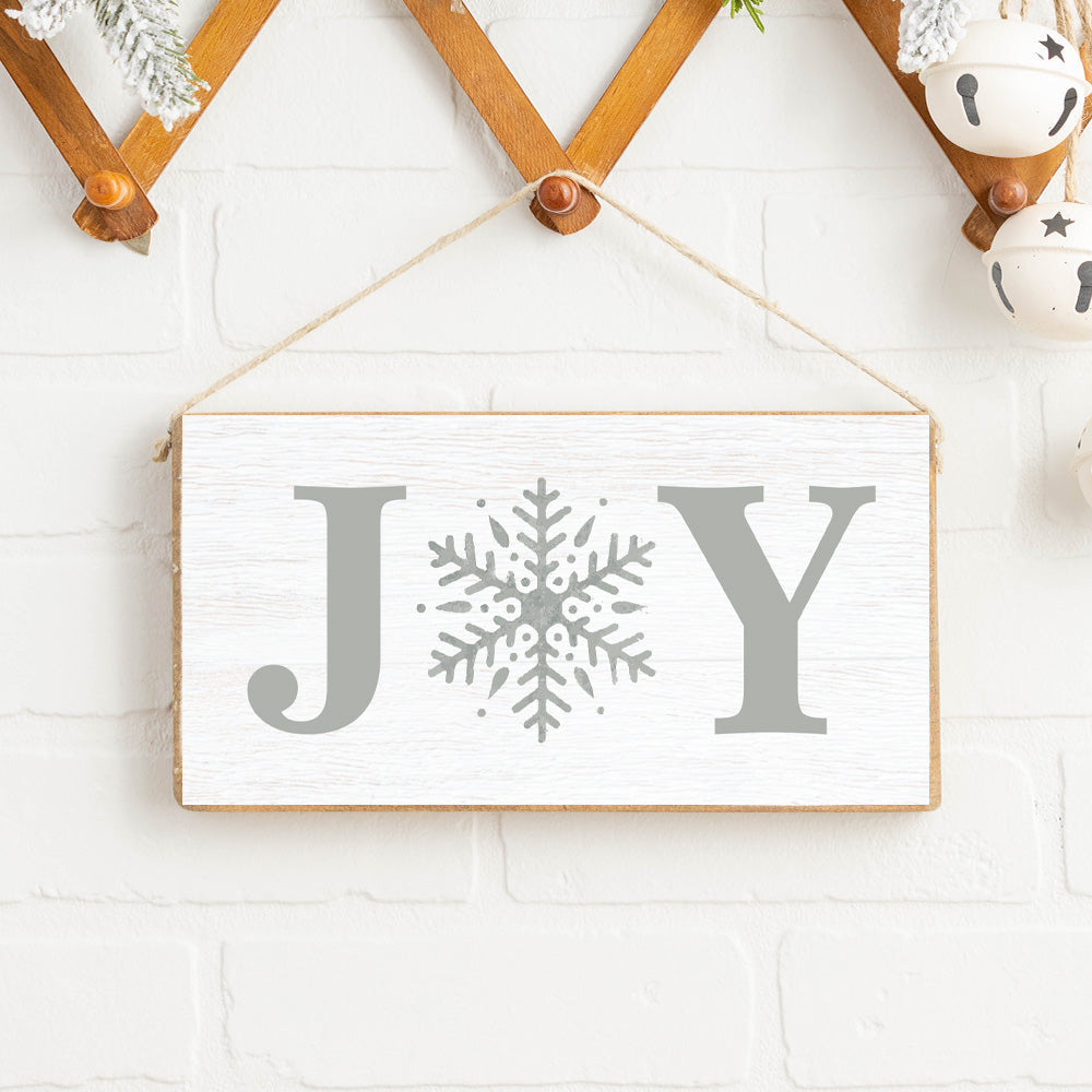 A rustic pine twine sign with the word 'JOY' and a snowflake design, hanging on a wall with a white brick background and decorative balls. Available at Be Charmed