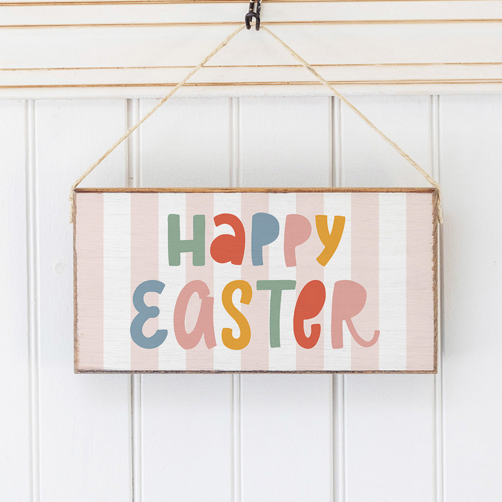 A wooden twine sign with a striped background and 'Happy Easter' written in colorful letters Hanging on a wall, attached to a rustic string. Available at Be Charmed