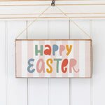 A wooden twine sign with a striped background and 'Happy Easter' written in colorful letters Hanging on a wall, attached to a rustic string. Available at Be Charmed