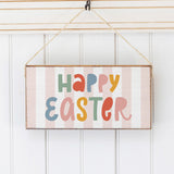 A wooden twine sign with a striped background and 'Happy Easter' written in colorful letters Hanging on a wall, attached to a rustic string. Available at Be Charmed