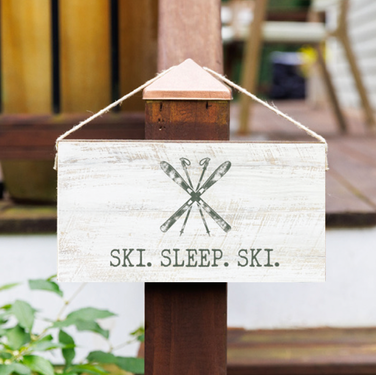 A wooden sign with a 'SKI, SLEEP, SKI' text and a graphic of skis, hanging on a wooden post outdoors. Available at Be Charmed