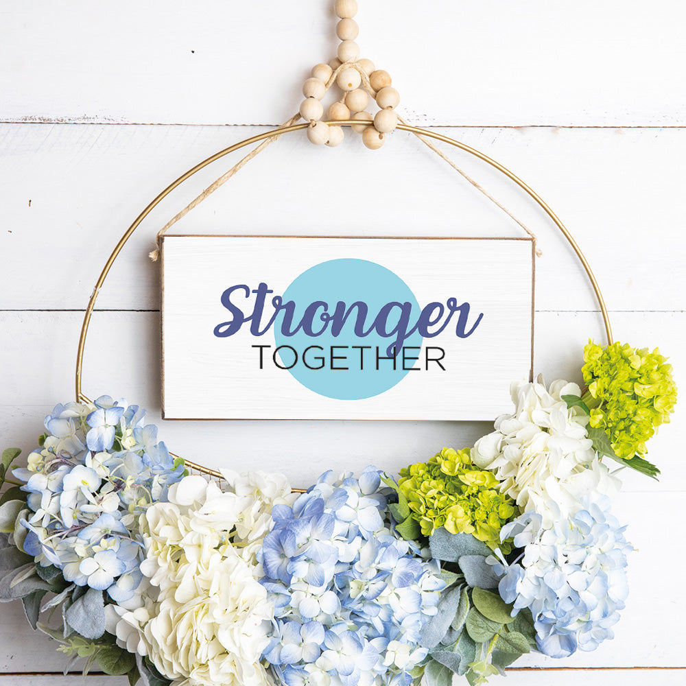 A rustic style wooden sign with a blue dot and the text 'STRONGER TOGETHER' displayed, surrounded by a twine frame and adorned with floral arrangements. Available at Be Charmed