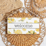 A round, handcrafted wooden sign with a twine border, featuring a watercolor print of lemons and the word 'WELCOME' in bold letters. Available at Be Charmed