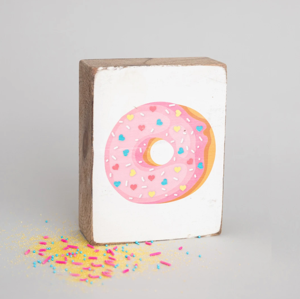 A rectangular wooden block with a painting of a donut with sprinkles on top, placed against a neutral background. Available at Be Charmed