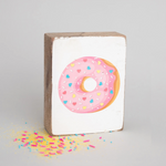 A rectangular wooden block with a painting of a donut with sprinkles on top, placed against a neutral background. Available at Be Charmed