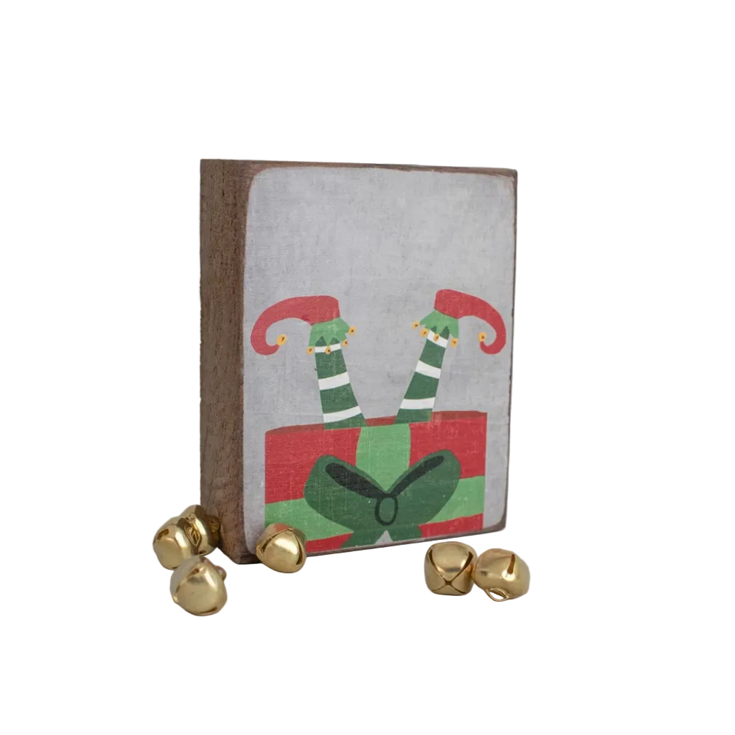 A decorative wooden block with a rustic design featuring an illustration of elf's legs in green and red with a white background, accompanied by gold-colored bells. Available at Be Charmed