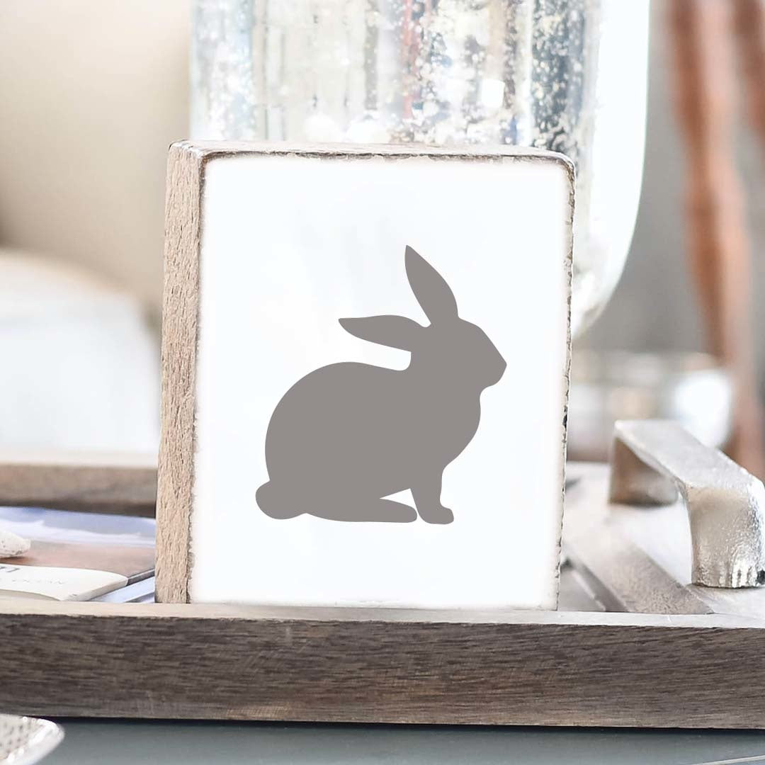 A gray wooden block decorative featuring a silhouette of a bunny on a tabletop. Available at Be Charmed