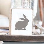 A gray wooden block decorative featuring a silhouette of a bunny on a tabletop. Available at Be Charmed