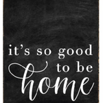 A rustic black wood block sign with the text 'It's So Good To Be Home' in white cursive font. Available at Be Charmed