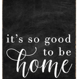A rustic black wood block sign with the text 'It's So Good To Be Home' in white cursive font. Available at Be Charmed