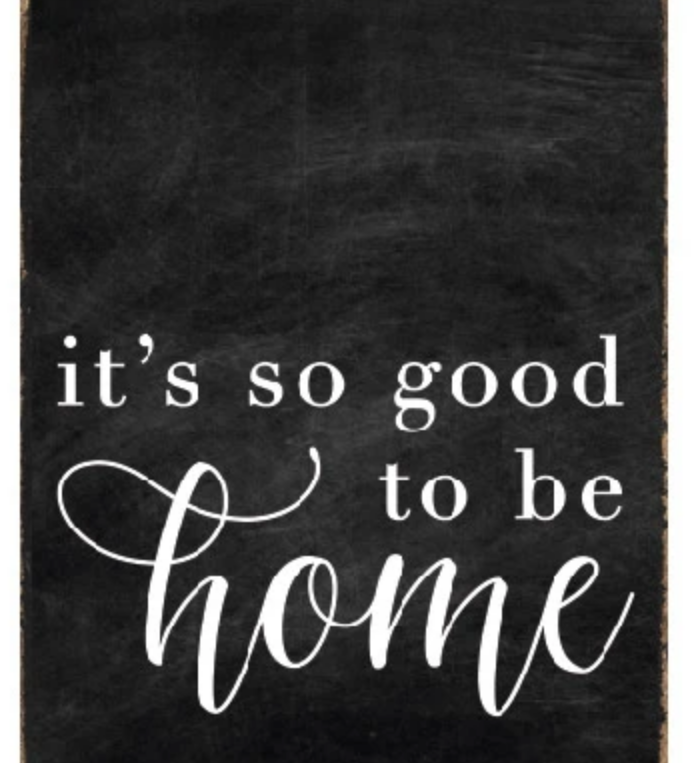 A rustic black wood block sign with the text 'It's So Good To Be Home' in white cursive font. Available at Be Charmed