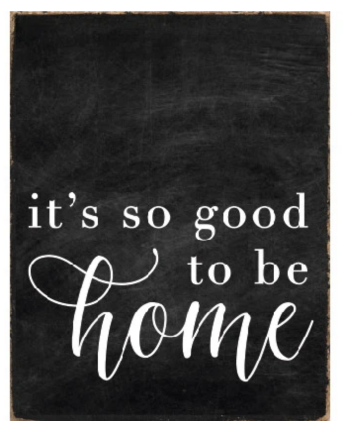 A rustic black wood block sign with the text 'It's So Good To Be Home' in white cursive font. Available at Be Charmed