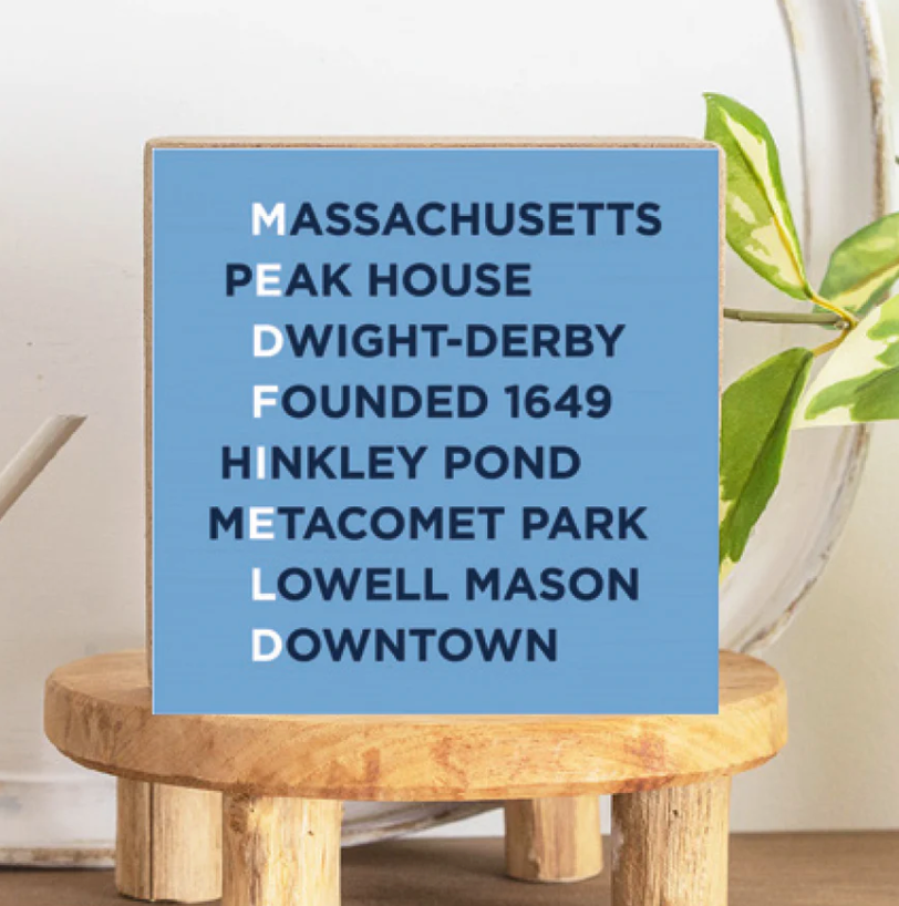 A decorative wood block sign with the text 'MEDFIELD' in capital letters and a list of locations in Medfield, Massachusetts, with a light blue background. The sign is placed on a wooden stand. Available at Be Charmed