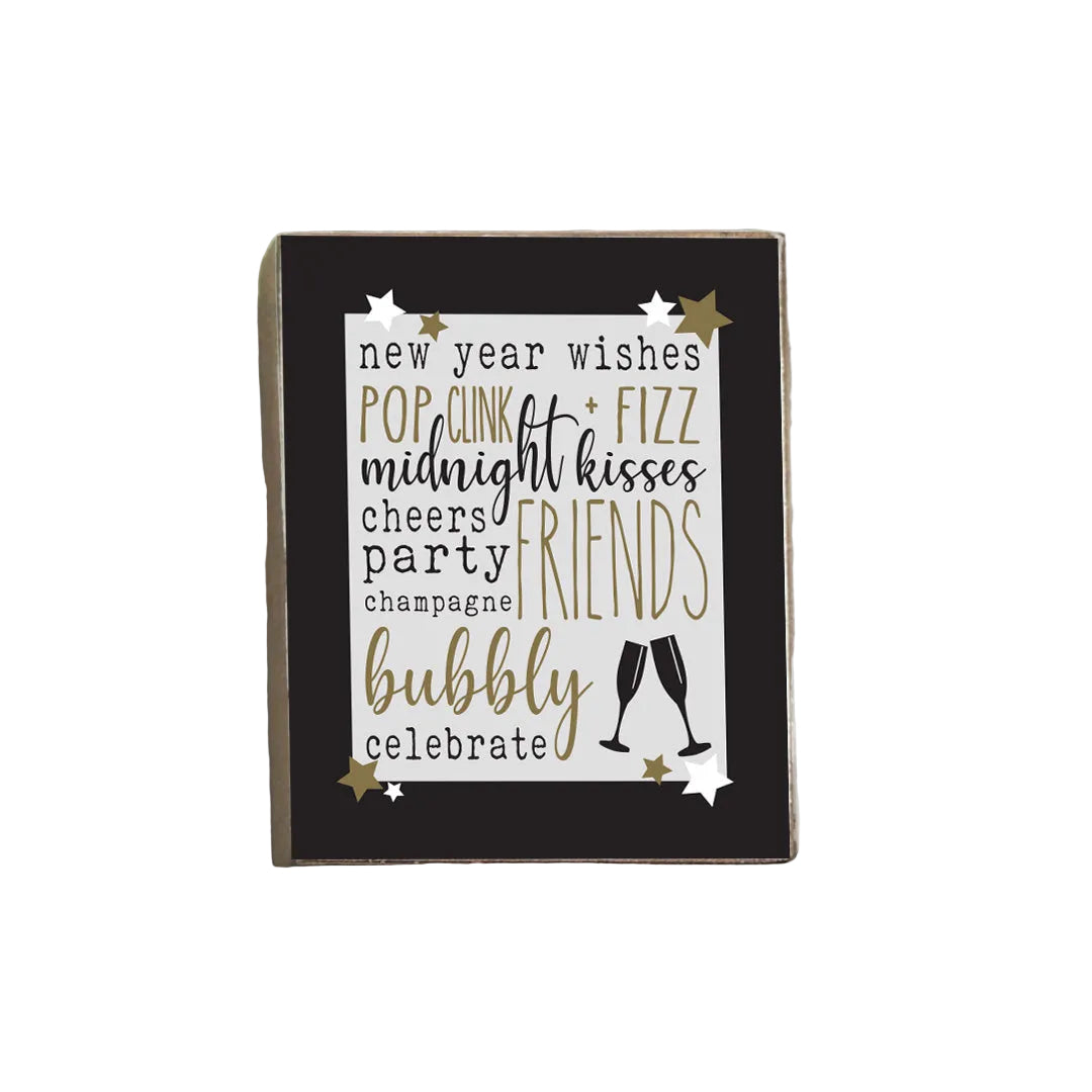 A decorative wooden block with a list of New Year's wishes written in a poetic format, accompanied by the word 'BUBBLY' at the bottom, indicating a celebratory tone. The block is dark in color with gold stars and a silver line drawing of a champagne bottle. Available at Be Charmed