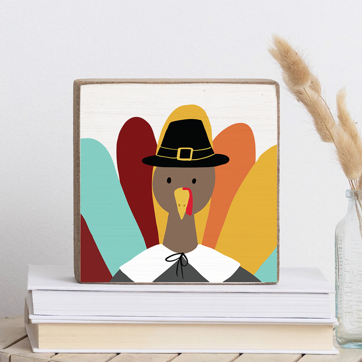 Decorative wooden block featuring a colorful illustration of a turkey in a Pilgrim hat and outfit, positioned on a shelf with books and a dry plant beside it. Available at Be Charmed