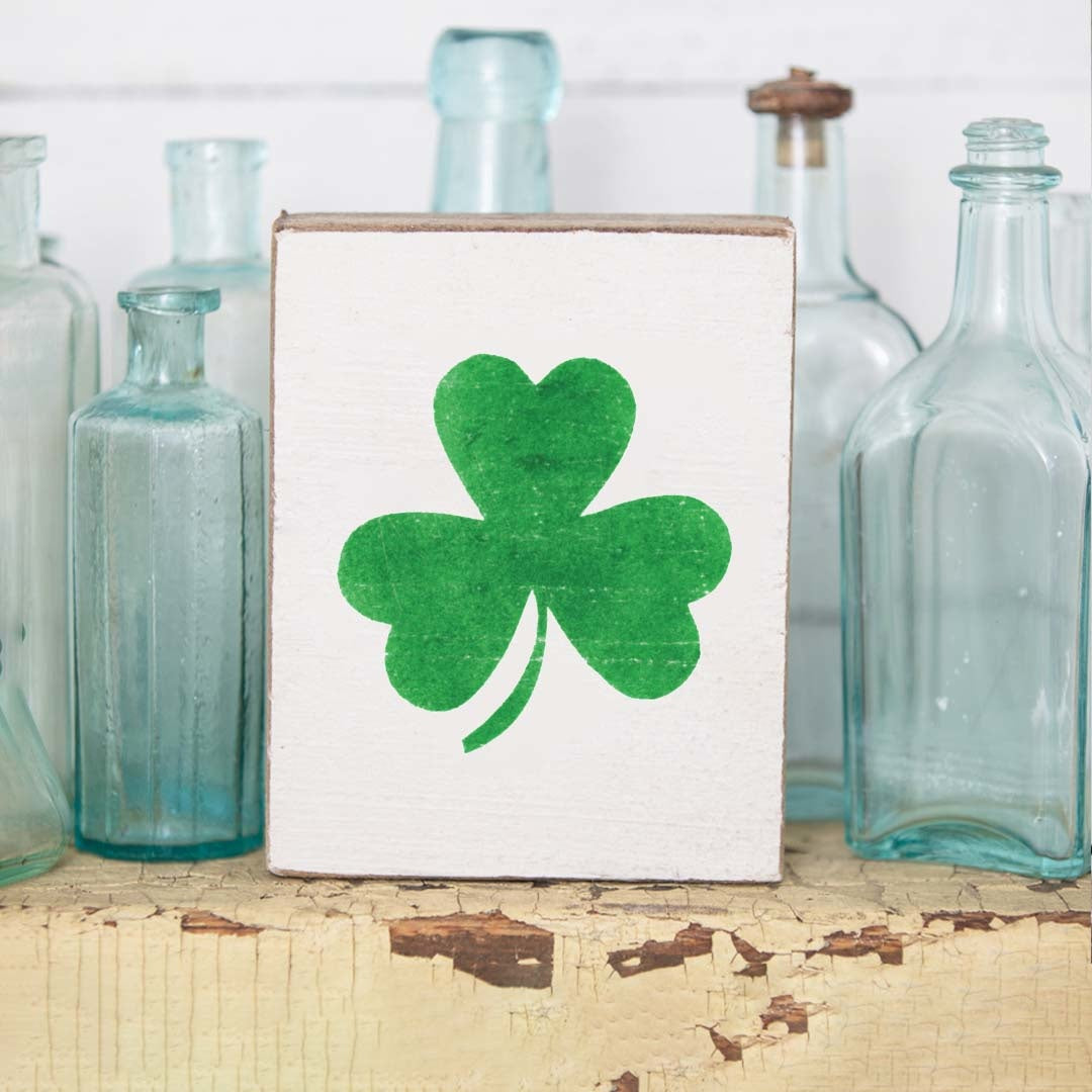 A rustic wooden block with a shamrock design on it, placed on a surface with glass bottles in the background. Available at Be Charmed