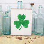 A rustic wooden block with a shamrock design on it, placed on a surface with glass bottles in the background. Available at Be Charmed