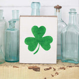 A rustic wooden block with a shamrock design on it, placed on a surface with glass bottles in the background. Available at Be Charmed