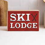 A decorative wooden block with a red background and the text 'SKI LODGE' in bold white letters, accompanied by a picture of skis. Available at Be Charmed