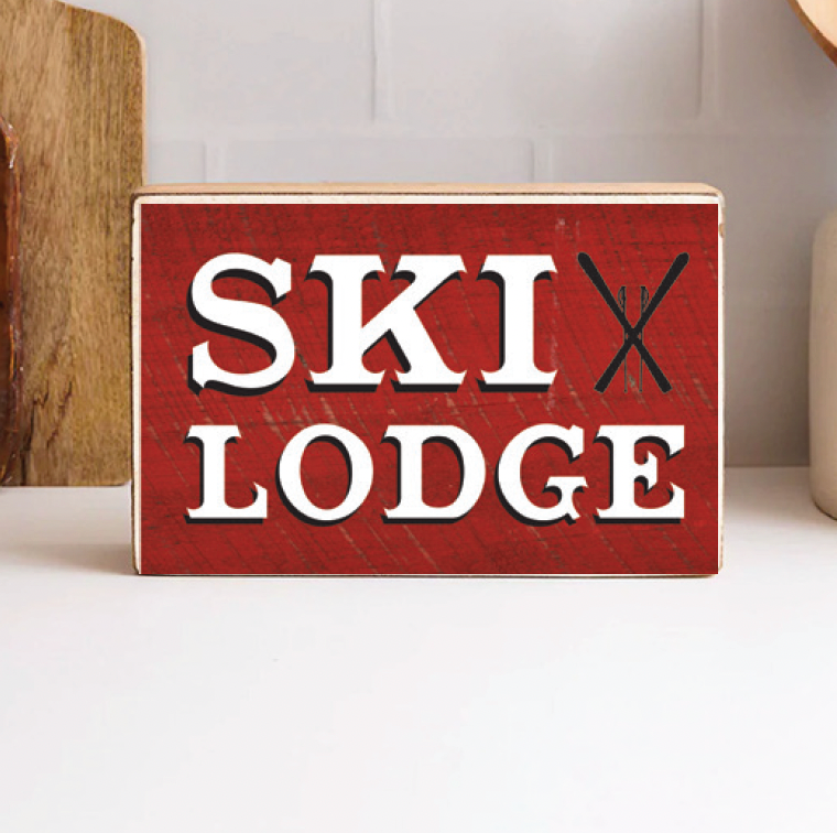 A decorative wooden block with a red background and the text 'SKI LODGE' in bold white letters, accompanied by a picture of skis. Available at Be Charmed
