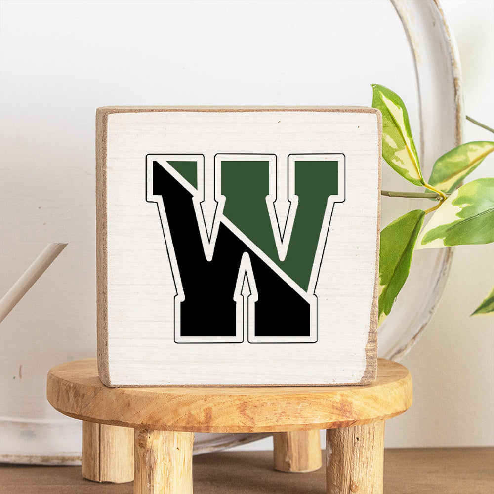 A handcrafted solid Rustic Marlin wooden block with a decorative letter 'W' in the center, placed on a wooden stool with a plant in the background. Available at Be Charmed