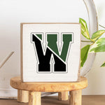 A handcrafted solid Rustic Marlin wooden block with a decorative letter 'W' in the center, placed on a wooden stool with a plant in the background. Available at Be Charmed