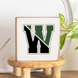A handcrafted solid Rustic Marlin wooden block with a decorative letter 'W' in the center, placed on a wooden stool with a plant in the background. Available at Be Charmed