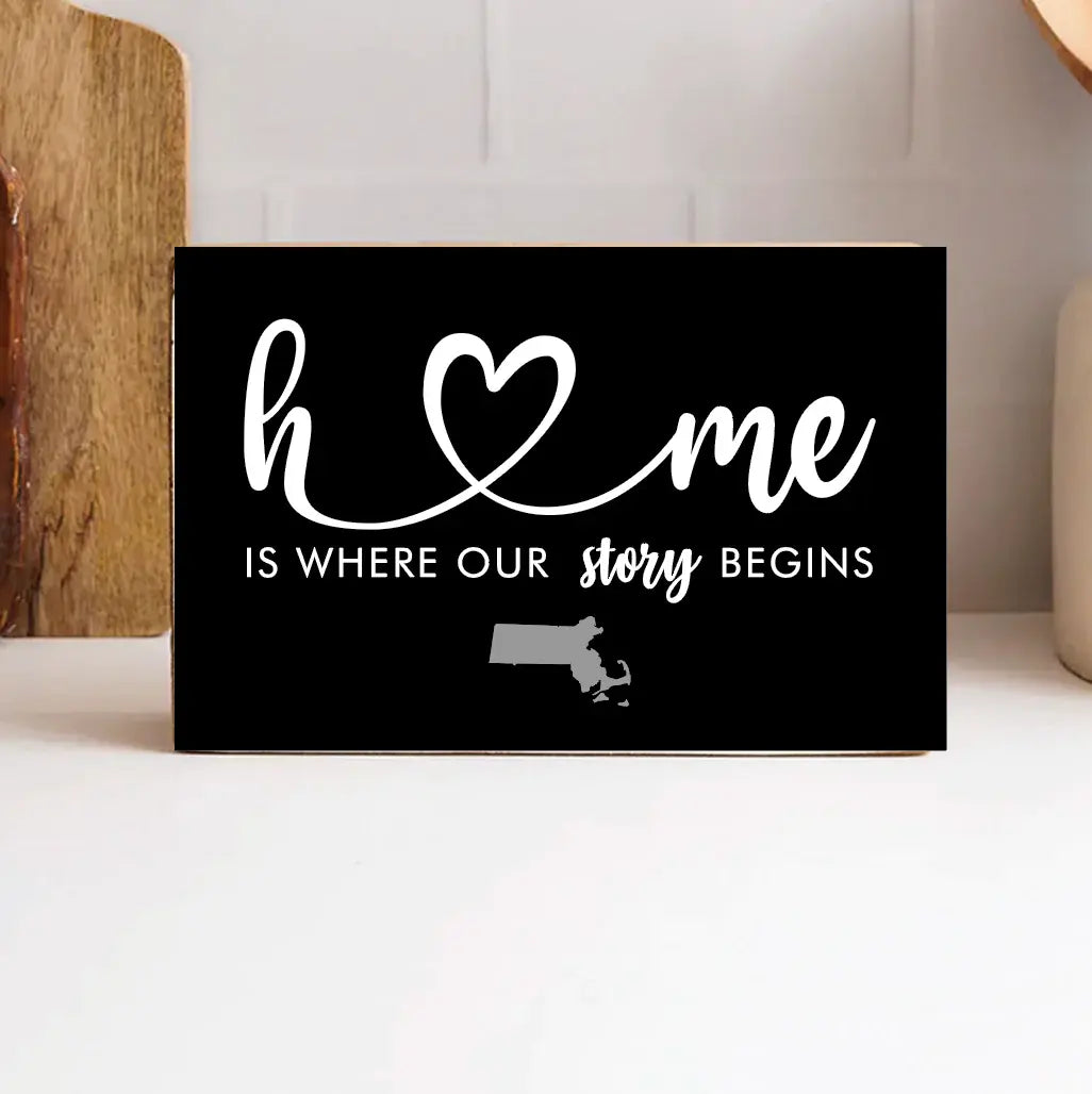 A black wooden block with white text and heart symbol, stating 'Home is where our story begins' with a small map outline representing Massachusetts. Available at Be Charmed