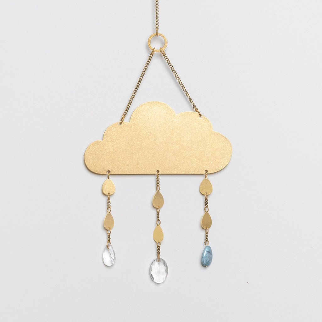 A brass suncatcher in the shape of a cloud with blue howlite gemstones and crystal pendants hanging from it. Available at Be Charmed