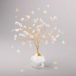 A suncatcher in the shape of a Tree of Life with gold branches and rose quartz stones, placed on a grey surface, with scattered rainbows around. Available at Be Charmed