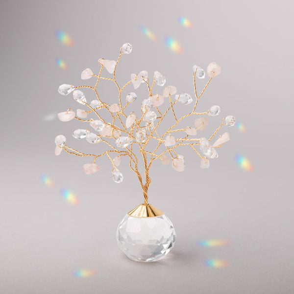 A suncatcher in the shape of a Tree of Life with gold branches and rose quartz stones, placed on a grey surface, with scattered rainbows around. Available at Be Charmed