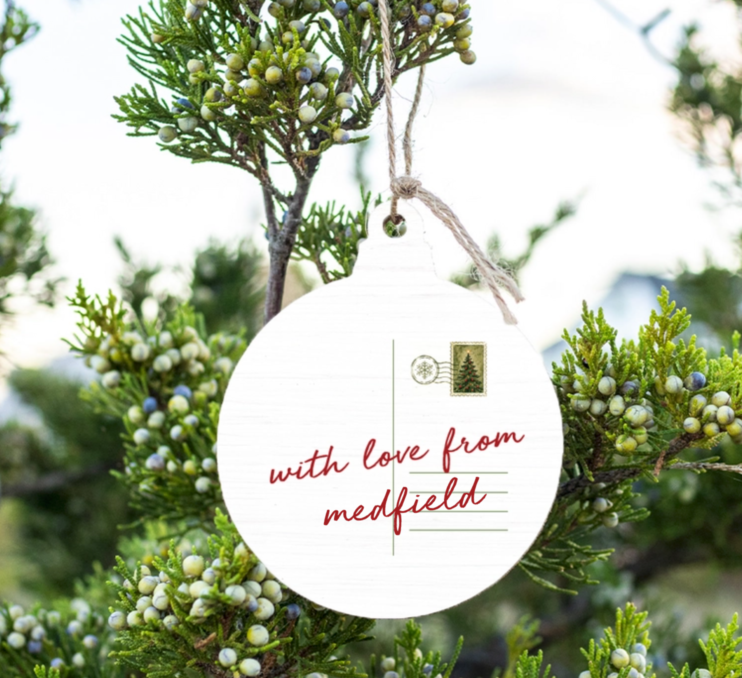 A Rustic Marlin round white hanging ornament with a postcard-style design and the phrase “with love from medfield” in red script, suspended by twine among green juniper branches with small berries. Available at Be Charmed
