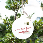 A Rustic Marlin round white hanging ornament with a postcard-style design and the phrase “with love from medfield” in red script, suspended by twine among green juniper branches with small berries. Available at Be Charmed