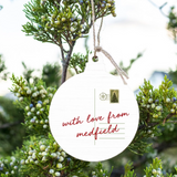 A Rustic Marlin round white hanging ornament with a postcard-style design and the phrase “with love from medfield” in red script, suspended by twine among green juniper branches with small berries. Available at Be Charmed