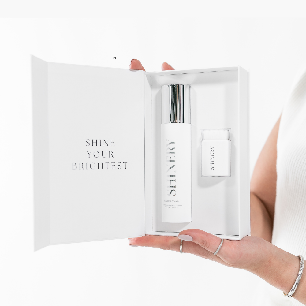 Hand holding a white box with skincare products and 'SHINE YOUR brightest' text. Available at Be Charmed
