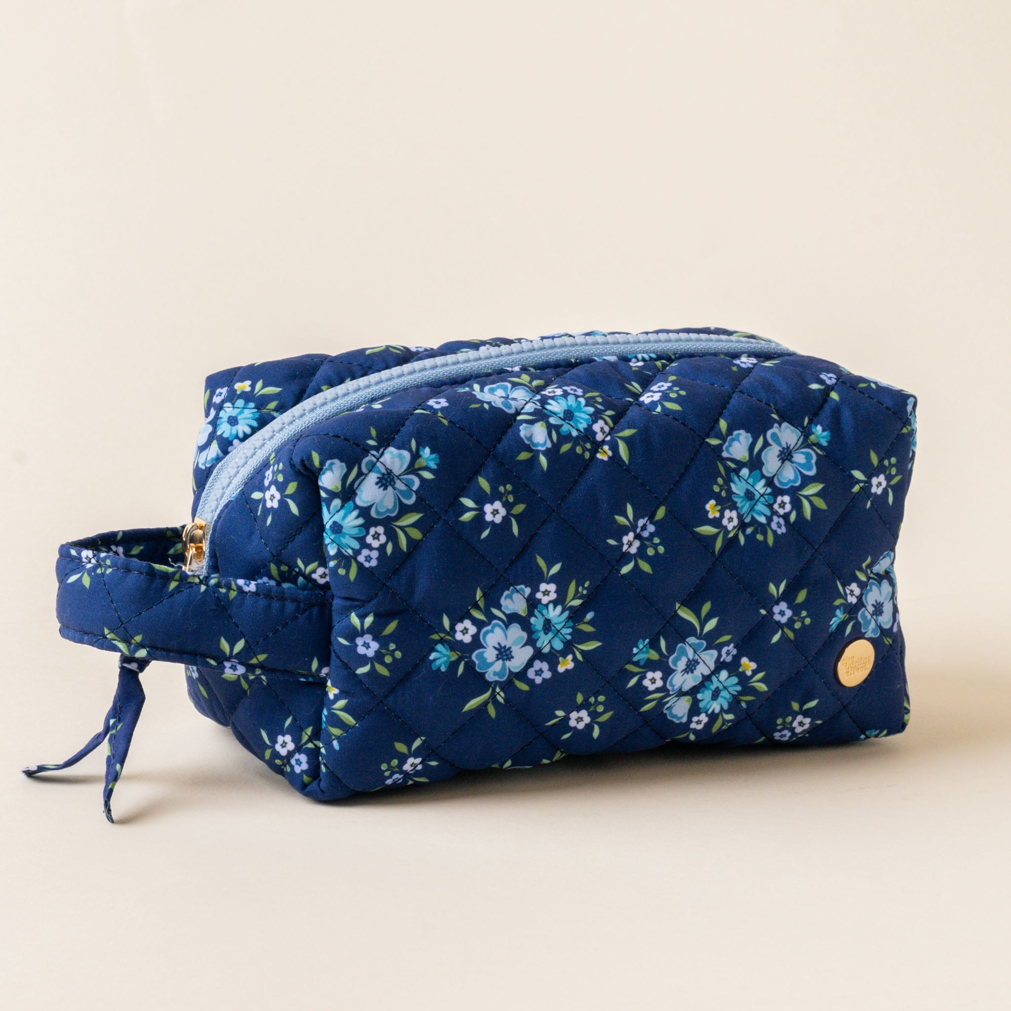 A quilted cosmetic pouch with a floral pattern, featuring a gold zipper and a carrying handle. Available at Be Charmed