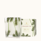 A clear glass candle with a heritage pine needle design, accompanied by a Thymes Frasier Fir candle box. Available at Be Charmed