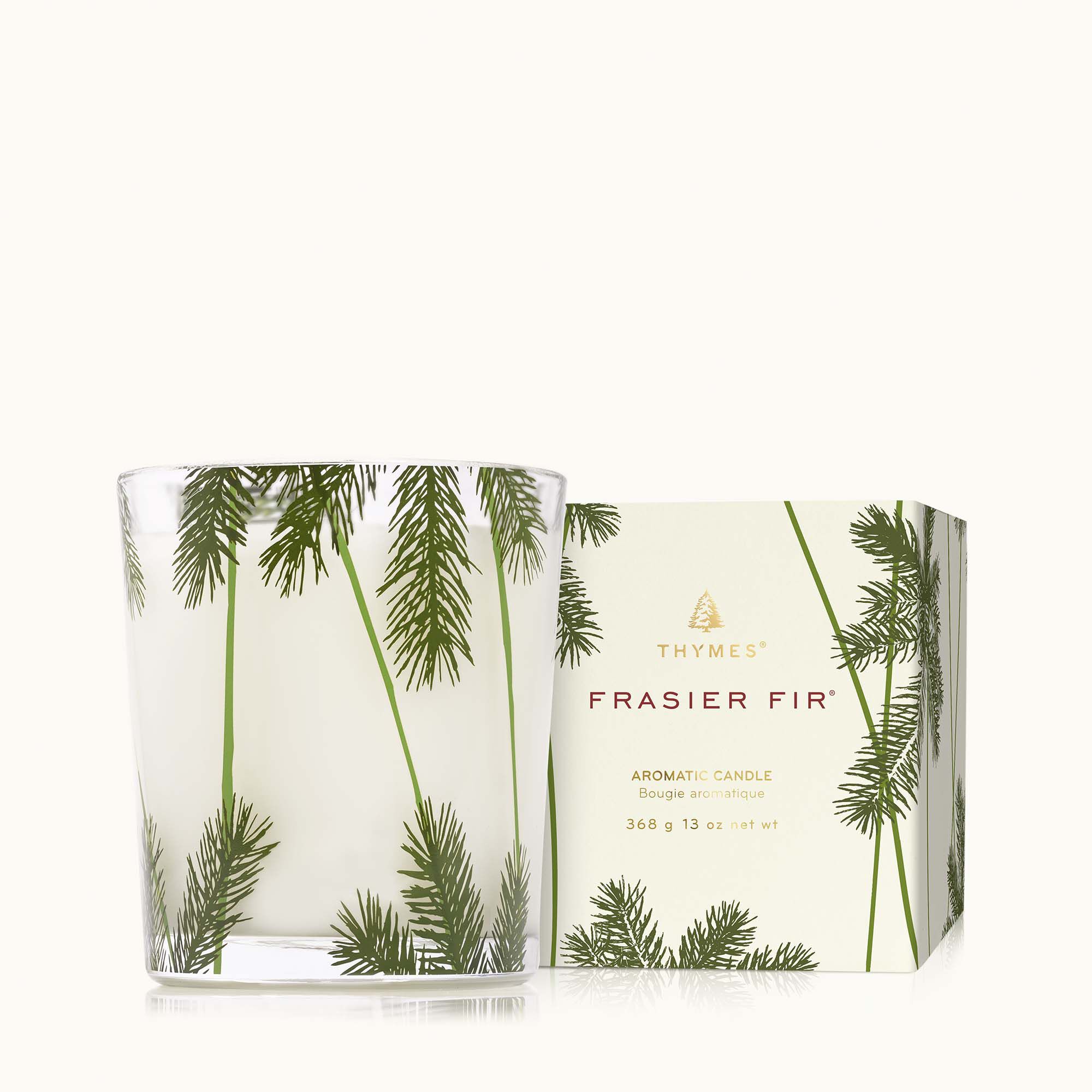 A clear glass candle with a heritage pine needle design, accompanied by a Thymes Frasier Fir candle box. Available at Be Charmed