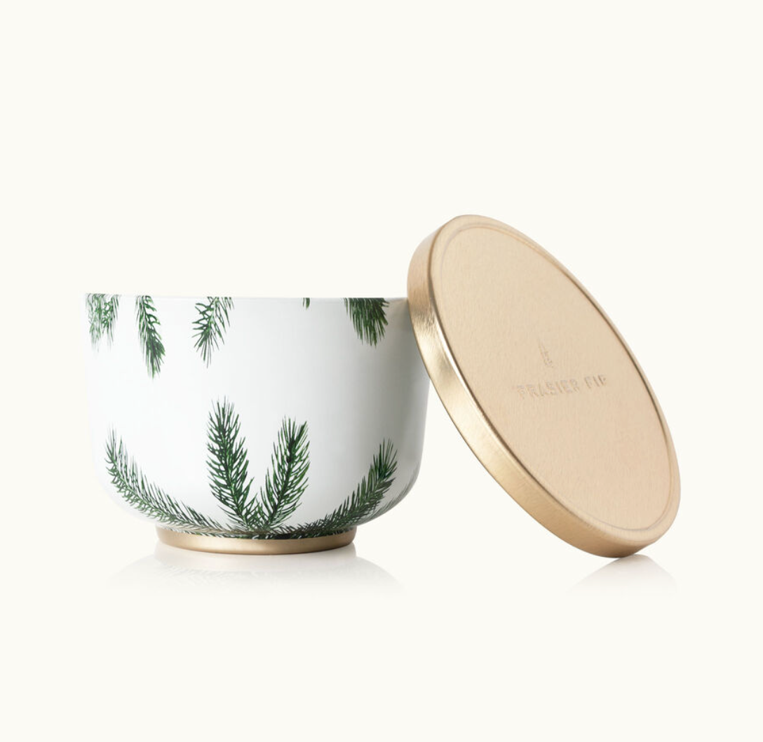 A gold-lidded candle tin with a white body and green Christmas tree patterns. The lid has a gold finish and the brand 'Thymes' is visible on it. Available at Be Charmed.  