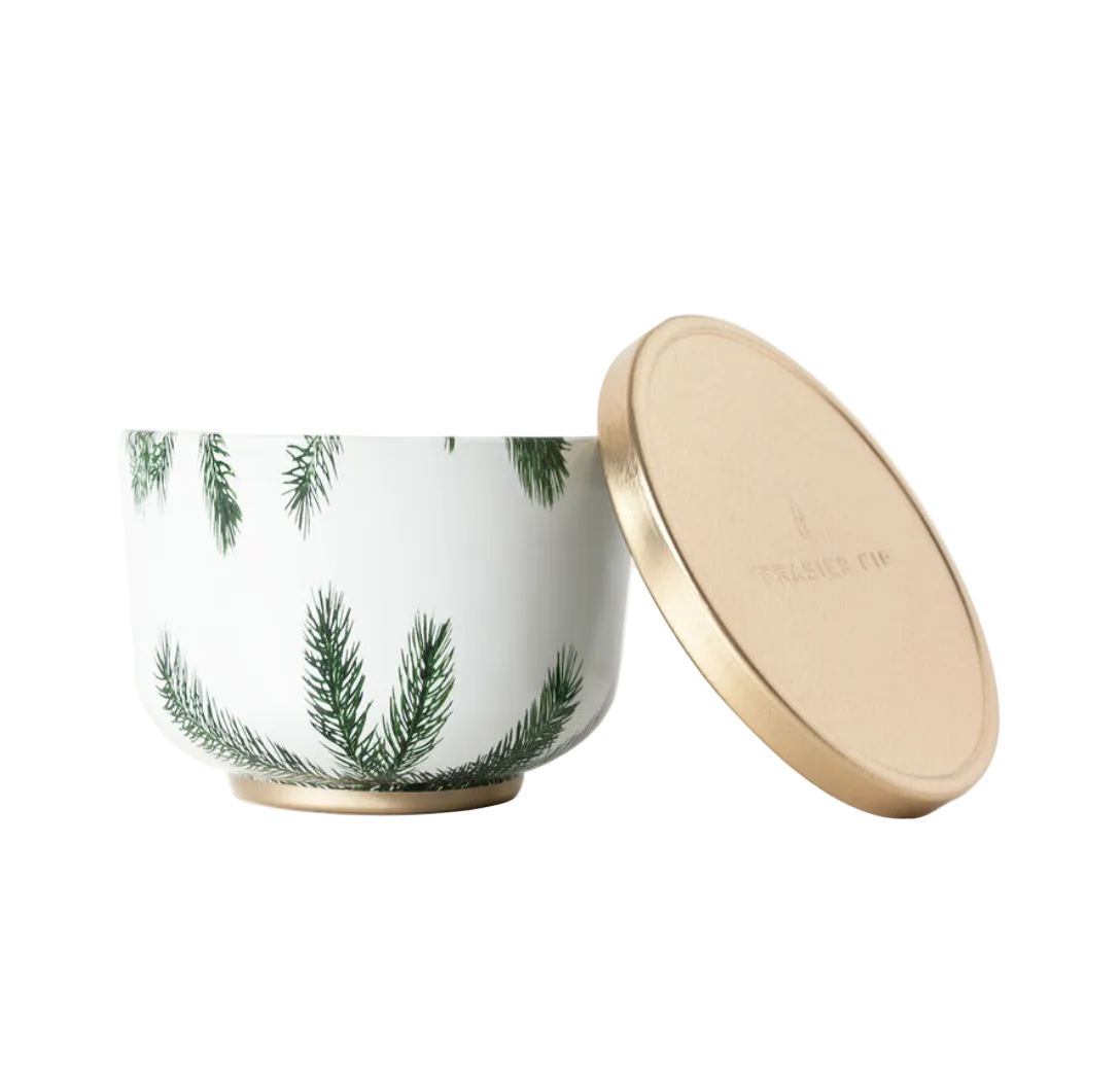 A gold-lidded candle tin with a white body and green Christmas tree patterns. The lid has a gold finish and the brand 'Thymes' is visible on it. Available at Be Charmed.  